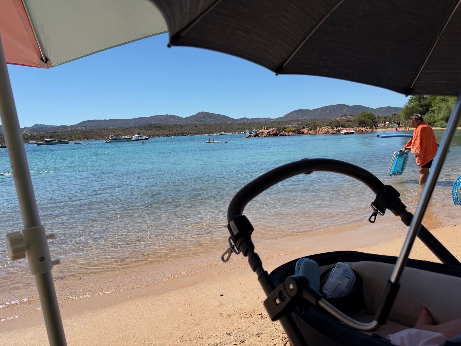 A pushchair under beach umbrellas beside calm shallow water